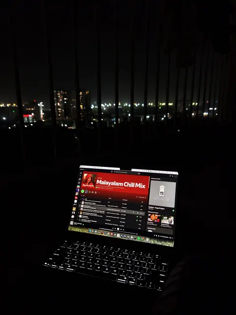 A laptop open in a dark room, its screen showing a music player queue, city lights out of focus behind it.