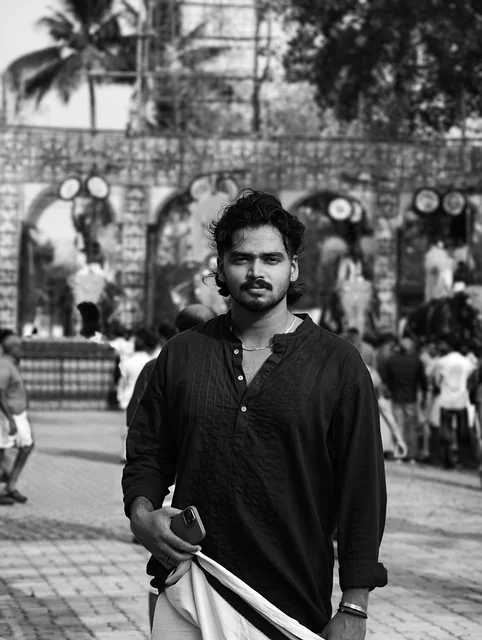 Black and white portrait of a man with curly hair and a moustache in a dark kurta and mundu, a crowded temple gateway soft behind him.