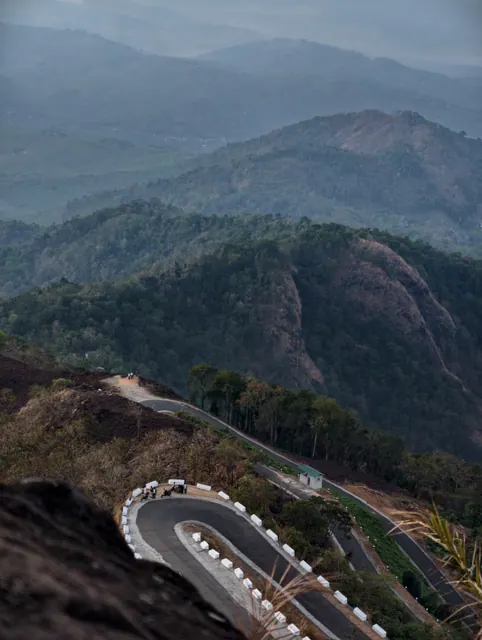 A ghat road folding back on itself in tight hairpin bends down a forested mountainside, seen from above in morning haze.