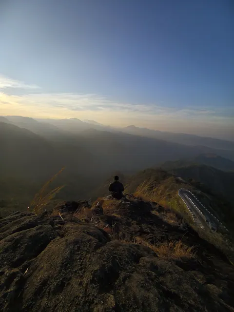 A dark mountain ridge running to the horizon at sunrise, with one small figure sitting on the highest rock.