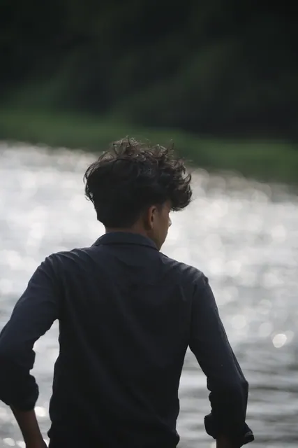 A person seen from behind in a dark shirt, standing waist-deep in a wide river with bright water and green banks beyond.
