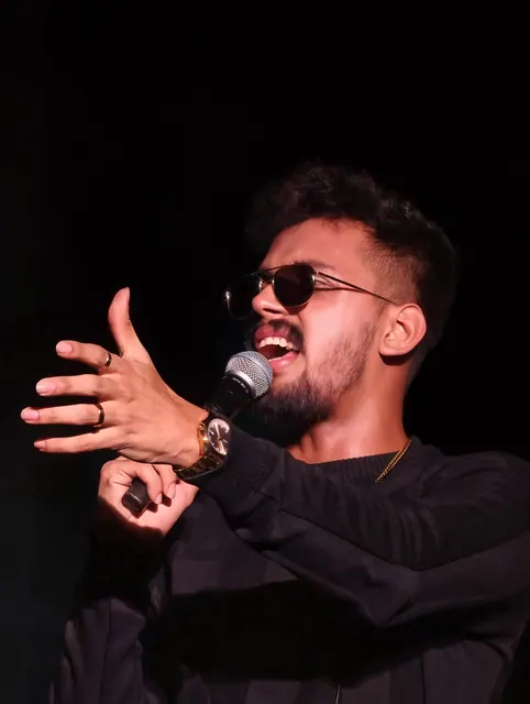A singer in black leans into a handheld microphone against a completely black stage, one hand open mid-gesture.