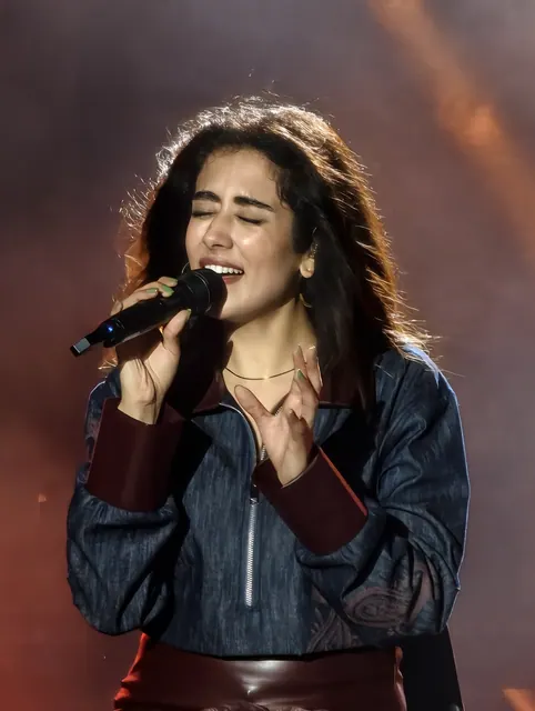 A singer in a denim jacket and leather belt performs under red and blue stage light, microphone raised, eyes closed.