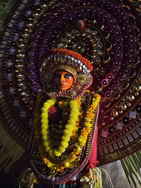 A Theyyam performer in a vast circular headdress of mirrors and beads, face painted, wearing heavy marigold garlands.